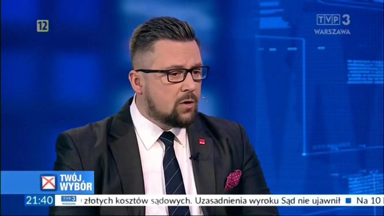 Mężczyzna w garniturze i okularach prowadzi rozmowę w studiu telewizyjnym, na tle niebieskiego ekranu z logiem TVP3 Warszawa i napisami na dole.