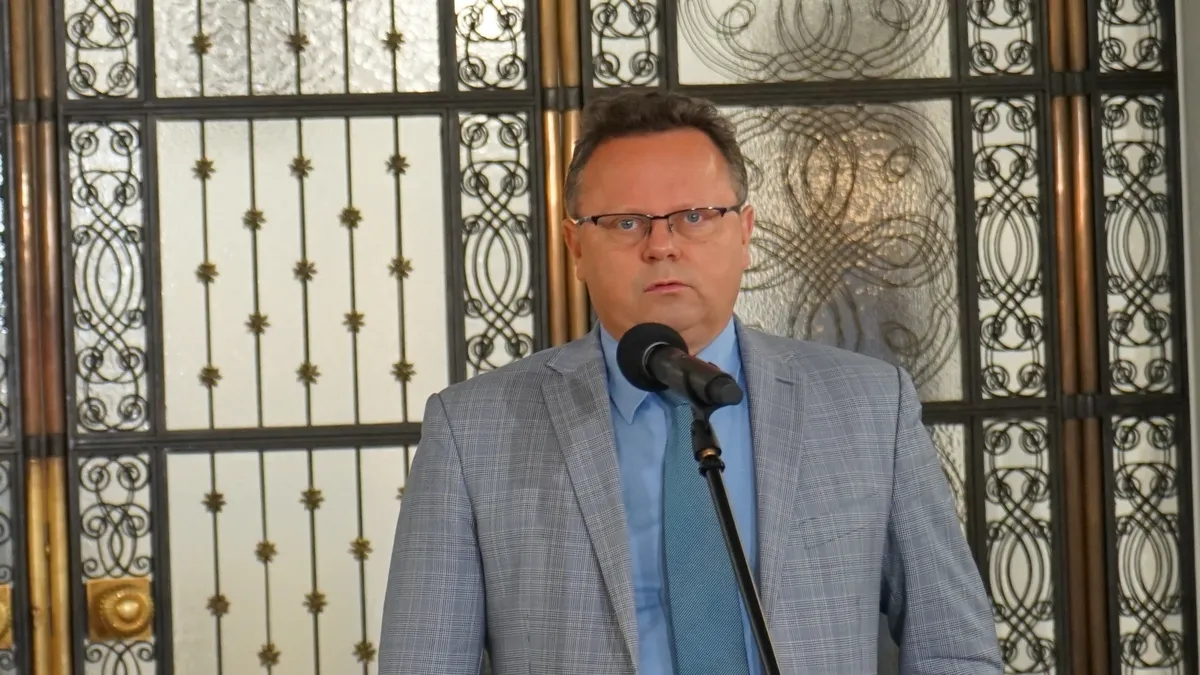 Mężczyzna w szarym garniturze w kratkę, niebieskiej koszuli i turkusowym krawacie stoi przy mikrofonie podczas konferencji prasowej, na tle ozdobnych metalowych paneli.