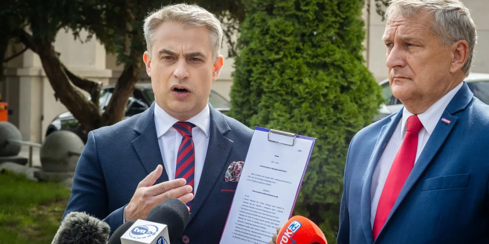 Dwóch mężczyzn w garniturach na zewnątrz budynku podczas konferencji prasowej; jeden z nich trzyma dokument i przemawia do mikrofonów.