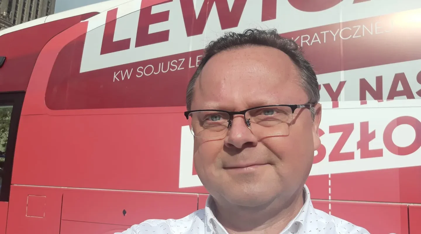 Mężczyzna w okularach przed czerwonym autobusem z napisem Lewica, robi selfie.