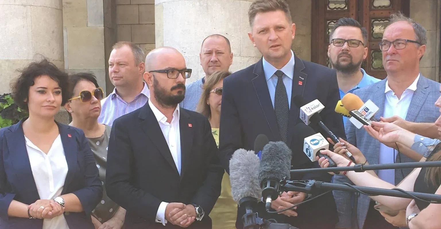 Grupa osób w garniturach udziela konferencji prasowej na zewnątrz, stojąc przed mikrofonami i dziennikarzami.