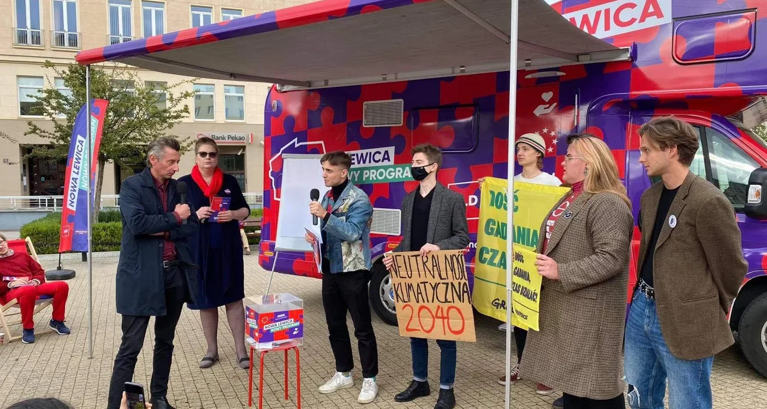 Kampania Nowej Lewicy na trasie: plenerowe wydarzenie z kamperem o jaskrawej fioletowo-czerwonej grafice, prowadzącym z mikrofonem i grupą uczestników trzymających plakaty.