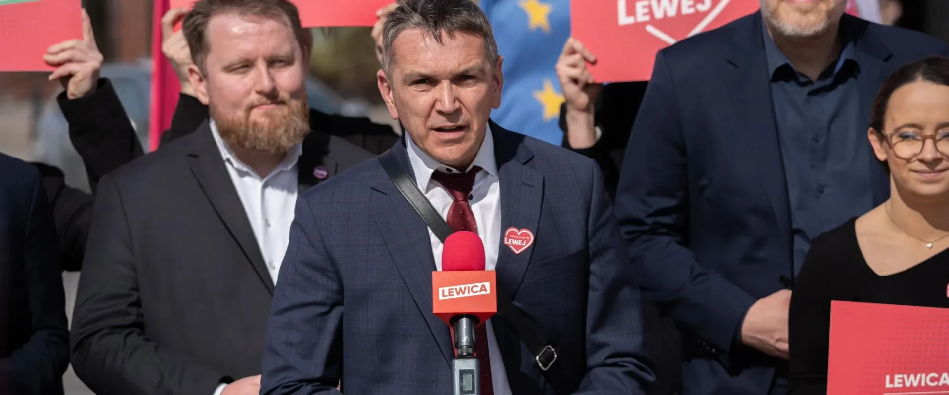 Grupa polityków Lewicy podczas konferencji prasowej, stojąca za mikrofonem z logiem Lewicy i trzymająca czerwone plakaty promujące darmowe posiłki dla uczniów.