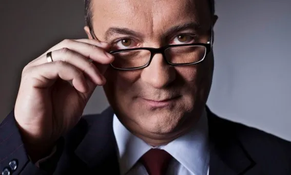 Mężczyzna w garniturze i krawacie, poprawiający okulary i patrzący poważnie w obiektyw na neutralnym tle.