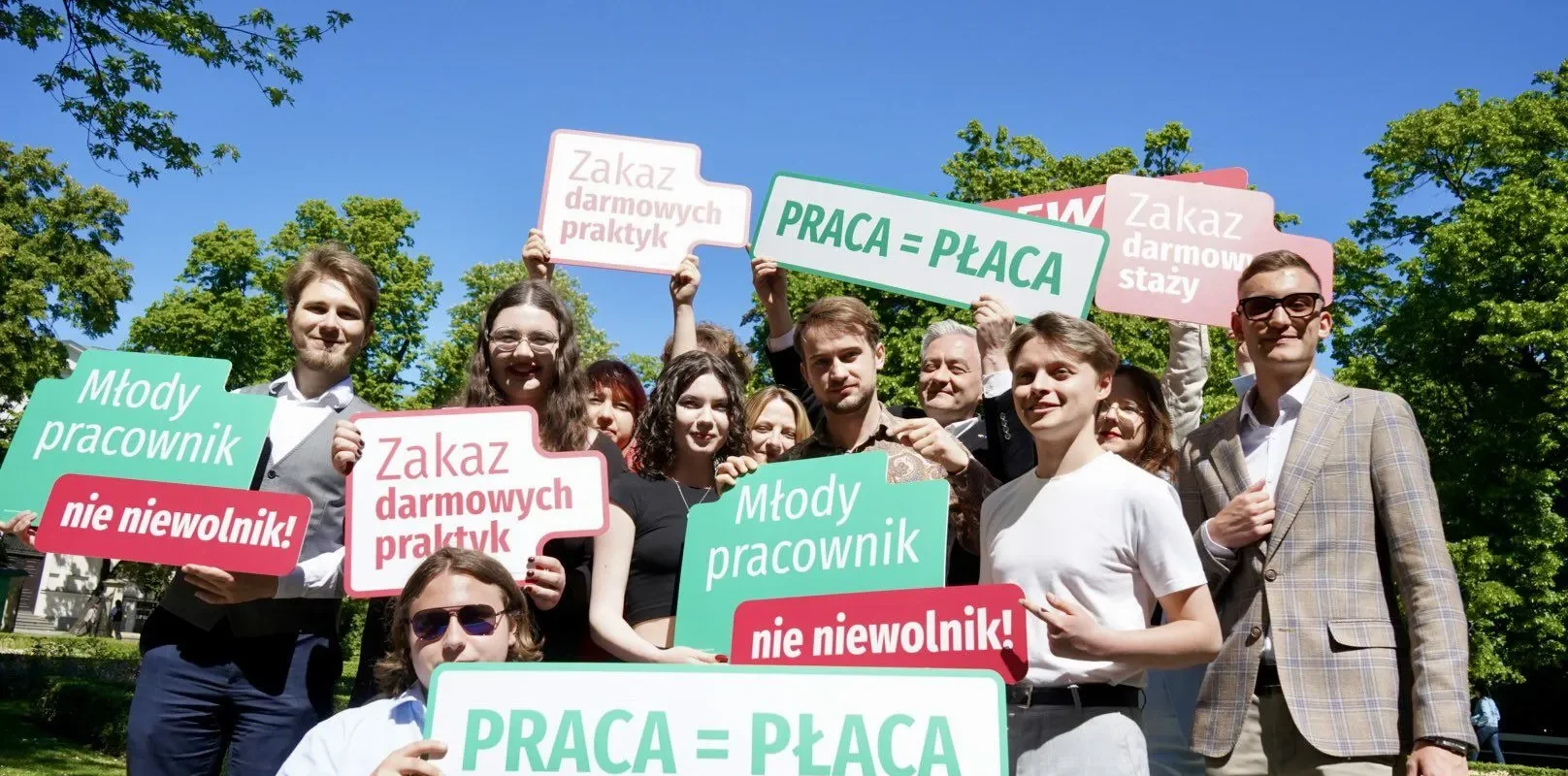 Grupa młodych ludzi stoi na zewnątrz i trzyma kolorowe plakaty z hasłami „PRACA = PŁACA” oraz „Zakaz darmowych praktyk”, protestując przeciw darmowym stażom.