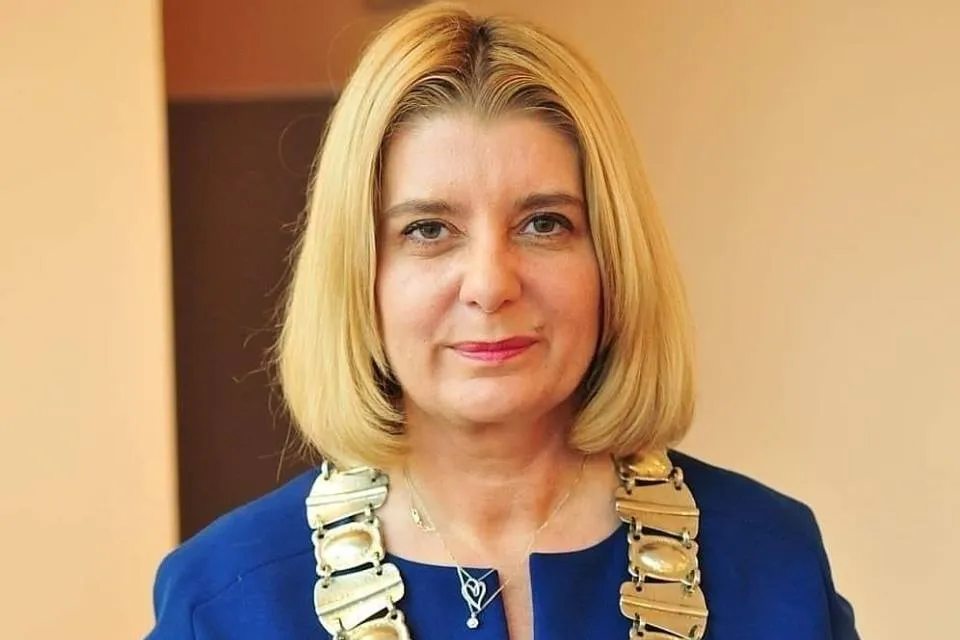 Portret kobiety o blond włosach, ubranej w granatowy żakiet i duży, błyszczący naszyjnik, na jasnym tle.