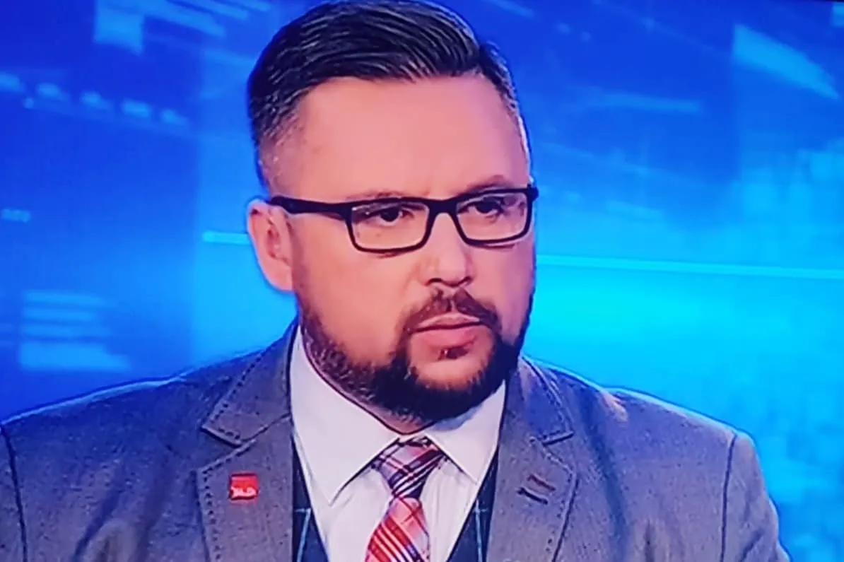 Mężczyzna w garniturze i okularach, siedzący w studiu telewizyjnym z niebieskim tłem.