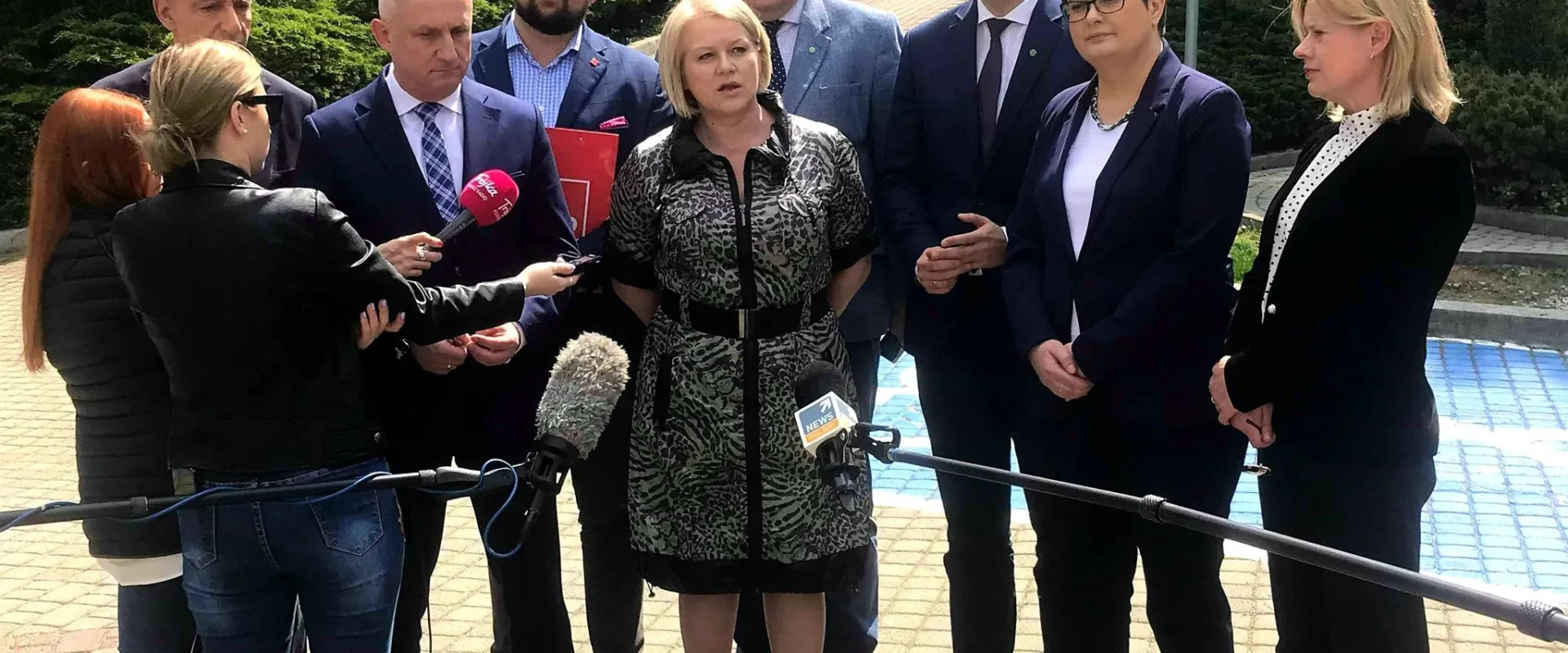 Grupa polityków i ekspertów w formalnych ubraniach udziela wywiadu dziennikarzom na zewnątrz budynku, stojąc w półokręgu obok mikrofonów.