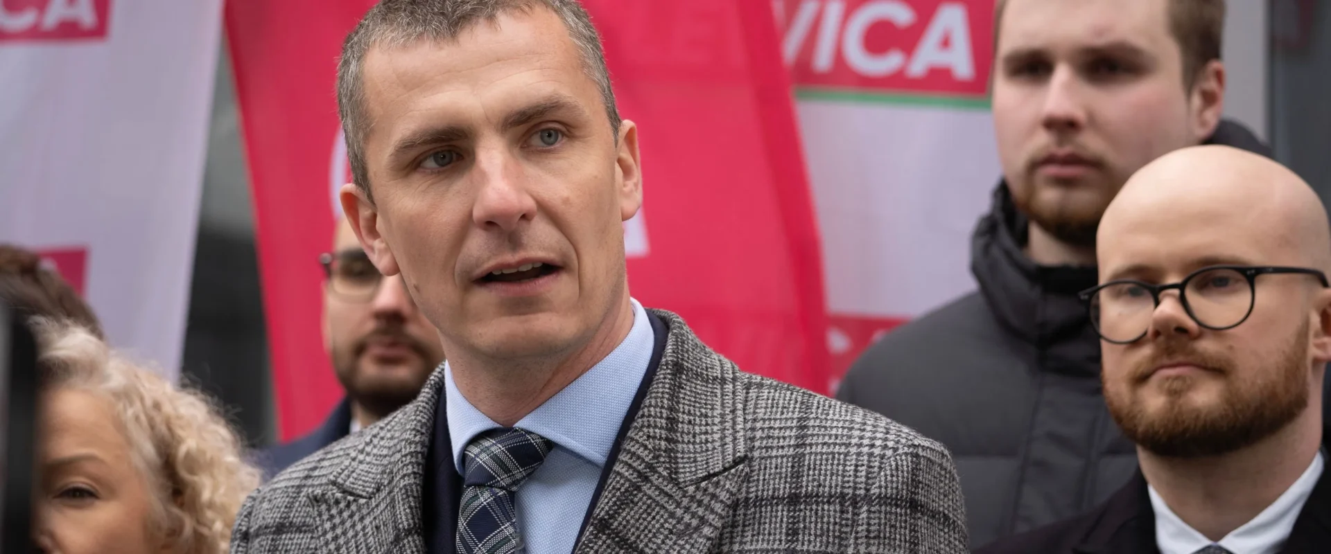 Mężczyzna w szarej kratkowanej marynarce i granatowym krawacie przemawia podczas wydarzenia politycznego na tle czerwono-białych flag.