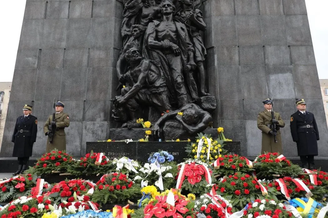 Na zdjęciu przedstawiono pomnik z figurami ludzi, złożonymi wieńcami kwiatów oraz strażników w mundurach, co wskazuje na uroczystość upamiętniającą ważne wydarzenie.