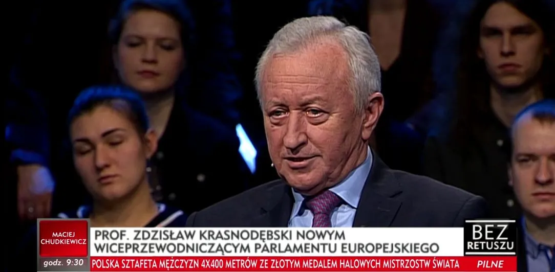 Starszy mężczyzna w garniturze i krawacie przemawia w studiu telewizyjnym, za nim widoczna publiczność i pasek z informacjami.