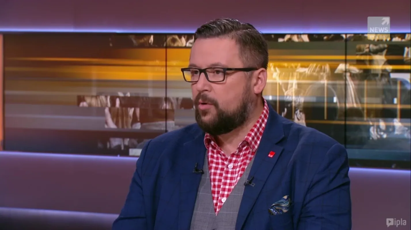 Mężczyzna w granatowym garniturze, czerwonej kraciastej koszuli i szarej kamizelce udziela wywiadu w studiu telewizyjnym.