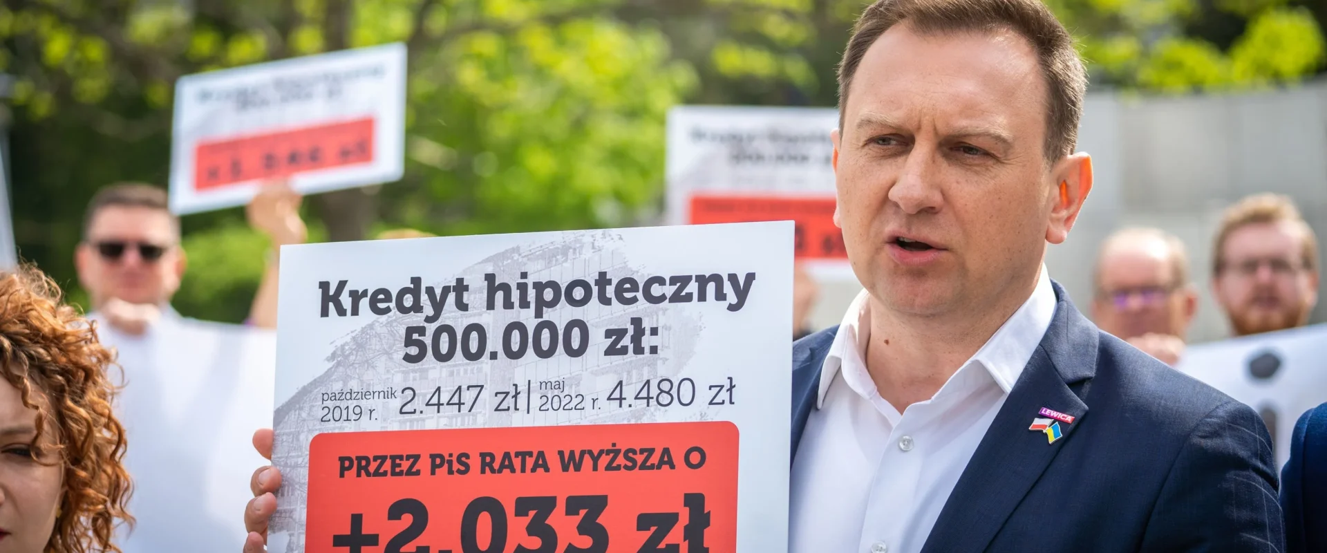 Mężczyzna w garniturze przemawia podczas demonstracji na zewnątrz, trzymając duży plakat o kredycie hipotecznym i rosnących ratach; w tle widoczni są inni uczestnicy i zielone drzewa.