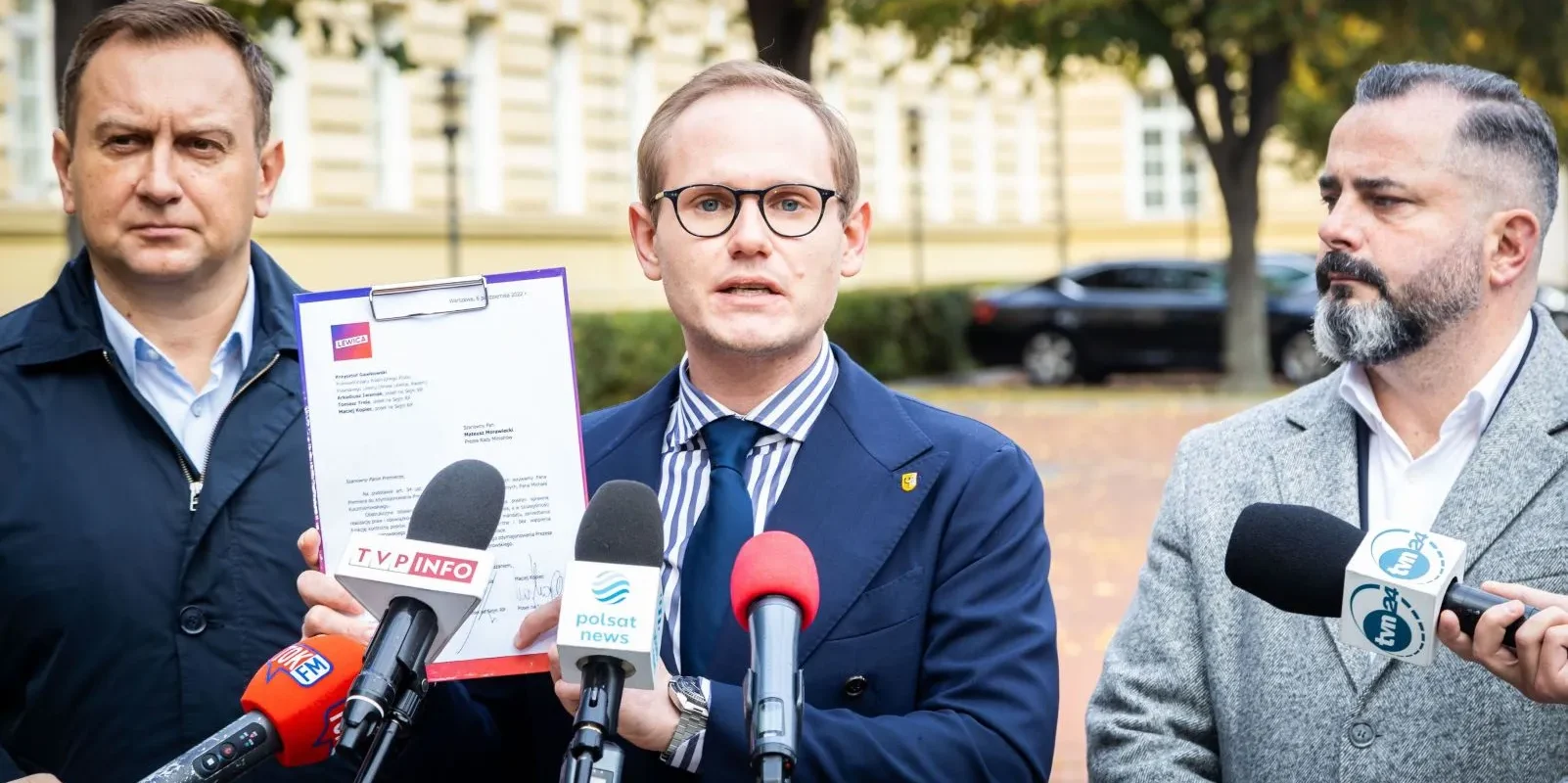 Na zdjęciu trzech mężczyzn stoi przed mikrofonami, z jednym z nich trzymającym dokument i przemawiającym do mediów.