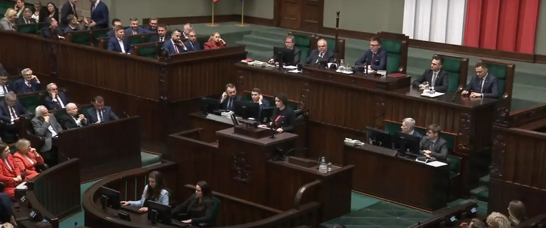 Parlamentarna sala plenarna wypełniona parlamentarzystami siedzącymi w rzędach, z centralnym pulpitem i stanowiskami z komputerami.