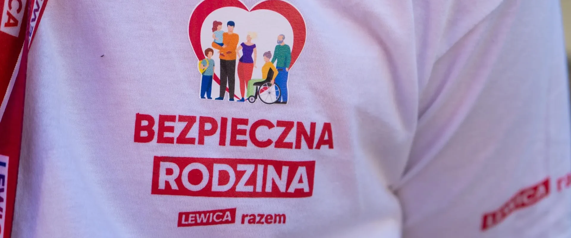 Na zdjęciu widoczna jest koszulka z napisem „BEZPIECZNA RODZINA” oraz grafiką przedstawiającą różnorodne postacie rodzinne, promującą inicjatywę „LEWICA razem”.