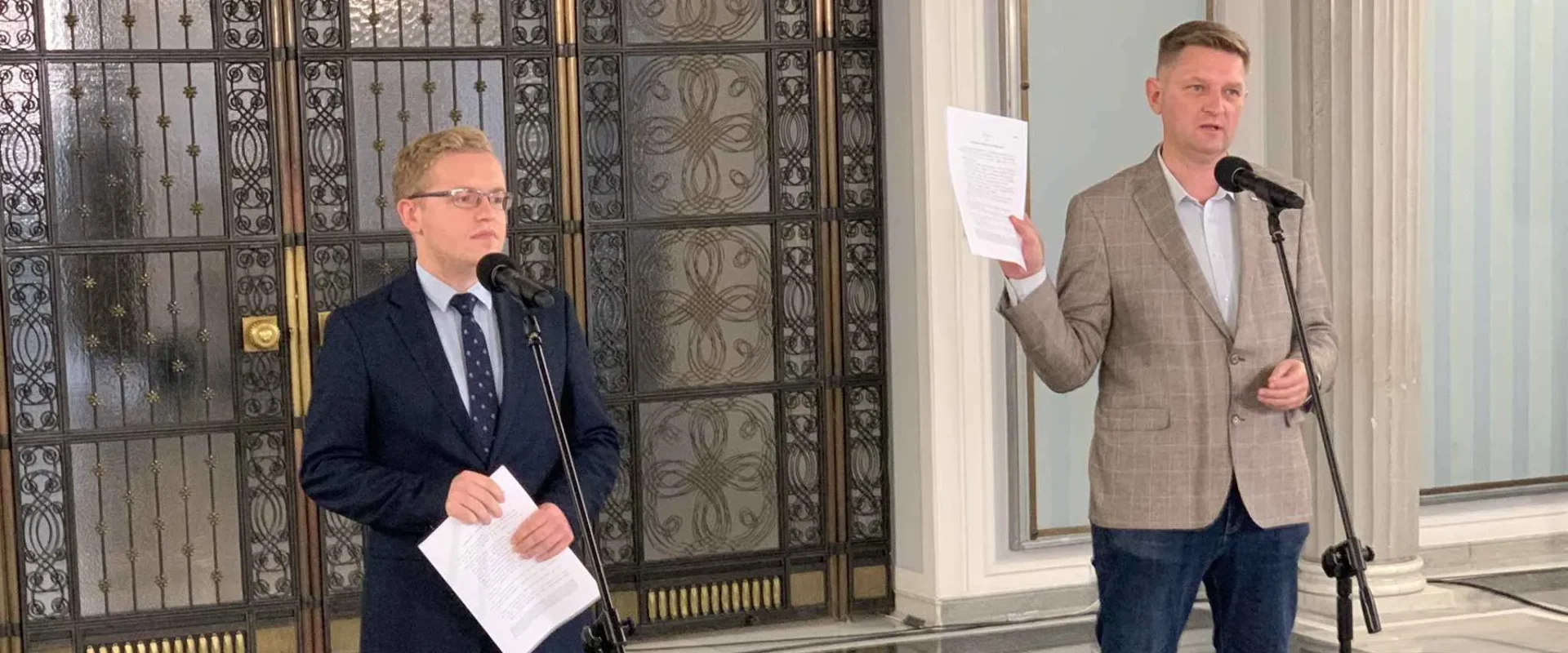 Dwóch mężczyzn w garniturach stoi przy mikrofonach w eleganckim wnętrzu, jeden trzyma kartki, a w tle widać ozdobne drzwi z kutymi panelami.