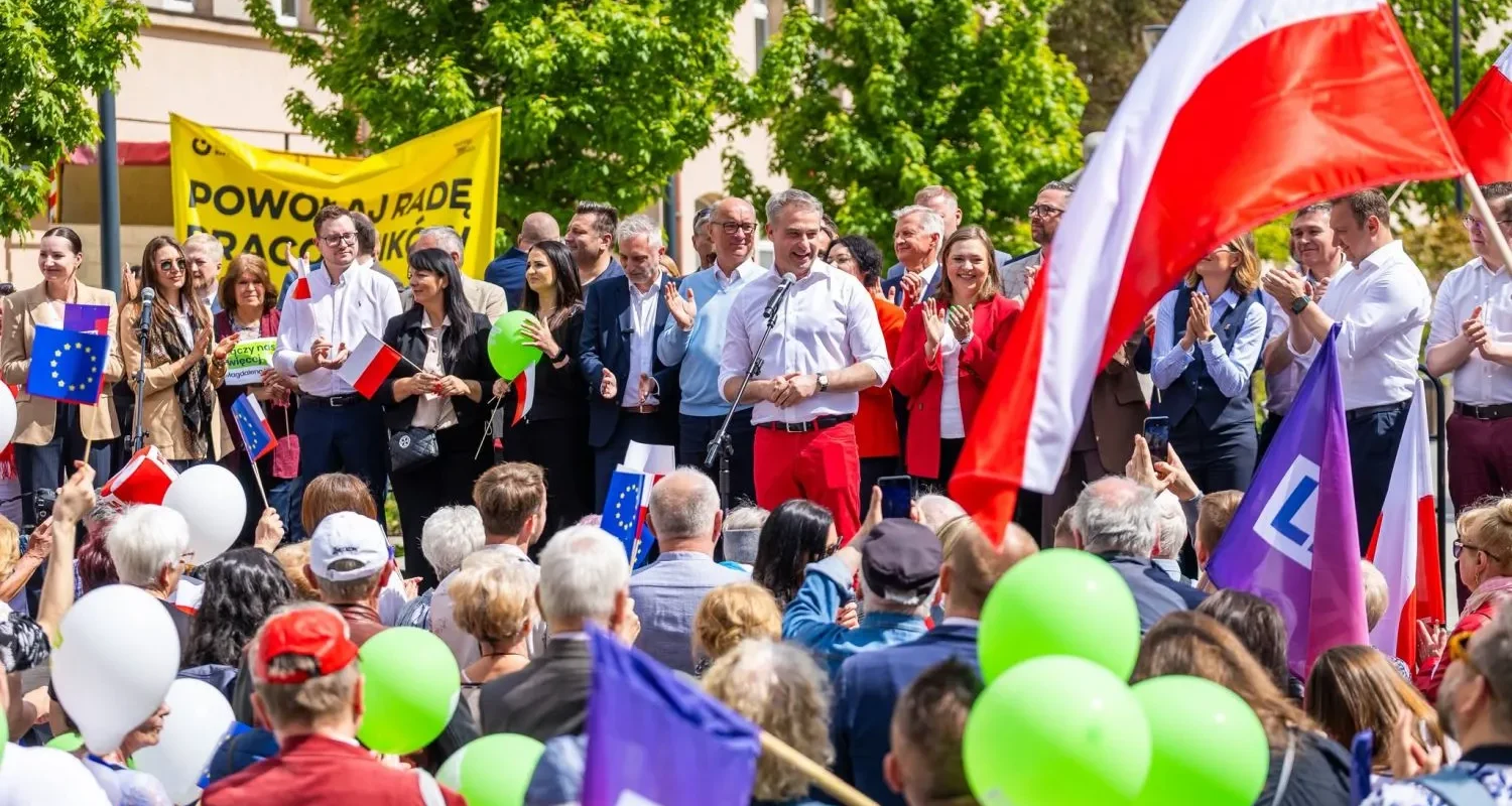 Grupa polityków stoi na scenie podczas wiecu wyborczego, przemawiając do zgromadzonego tłumu z flagami Polski i Unii Europejskiej oraz balonami.
