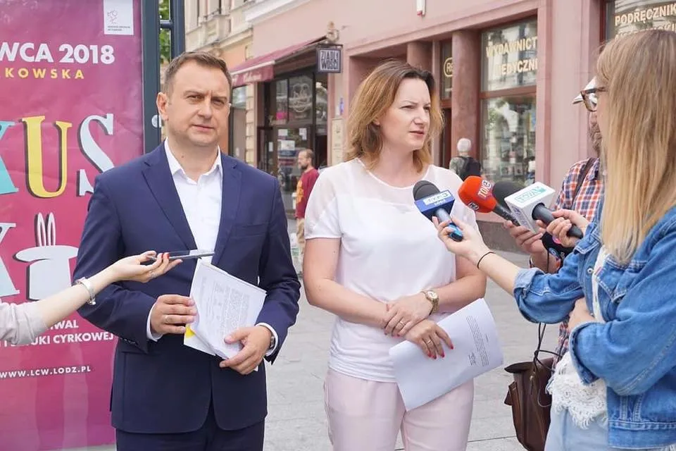 Na ulicy trwa konferencja prasowa dotycząca listu otwartego do Prezesa Kaczyńskiego: mężczyzna w granatowym garniturze trzyma dokumenty, obok niego stoi kobieta w białej bluzce, a reporterzy z mikrofonami prowadzą wywiad.
