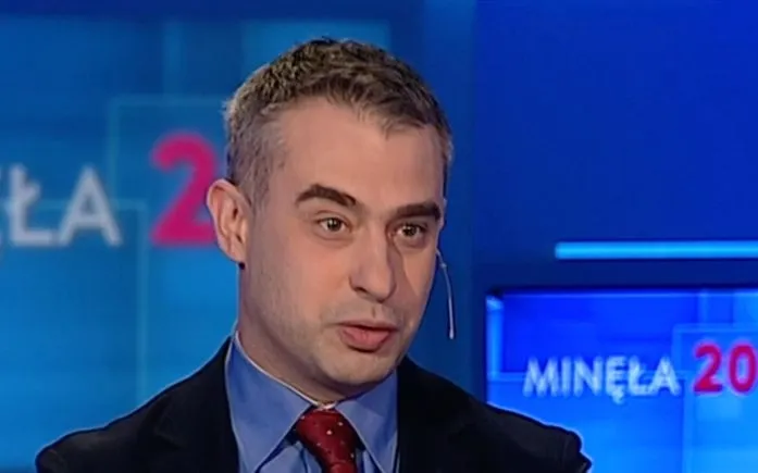 Mężczyzna w garniturze i czerwonym krawacie z mikrofonem nasadnym, mówiący w studiu telewizyjnym na niebieskim tle z widocznym napisem programu.