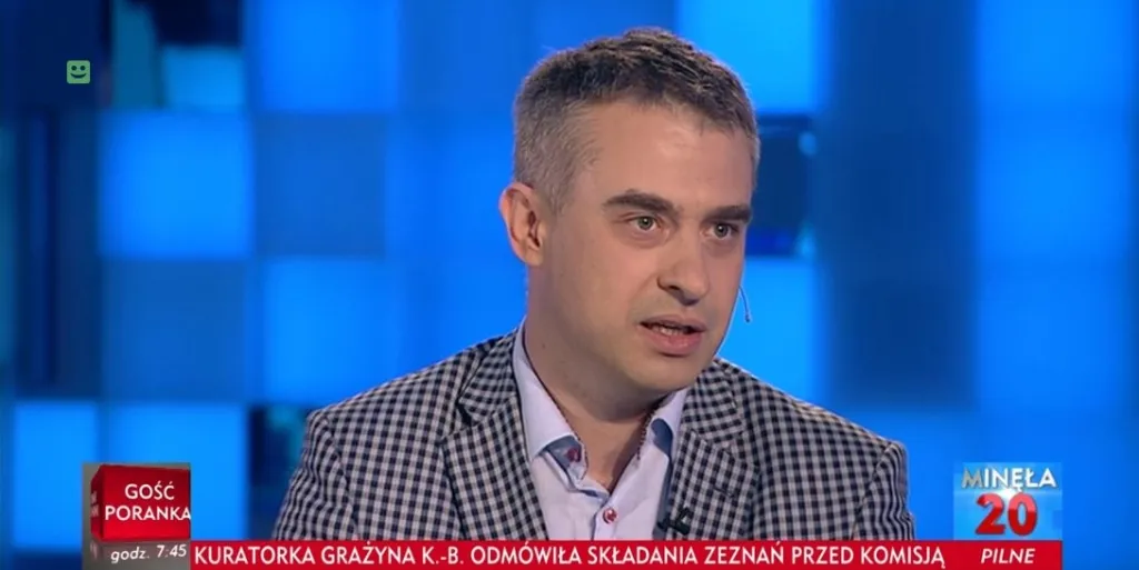 Mężczyzna w marynarce w kratkę z mikrofonem przy uchu mówi w studiu telewizyjnym na niebieskim tle, obok widoczny pasek informacyjny programu.
