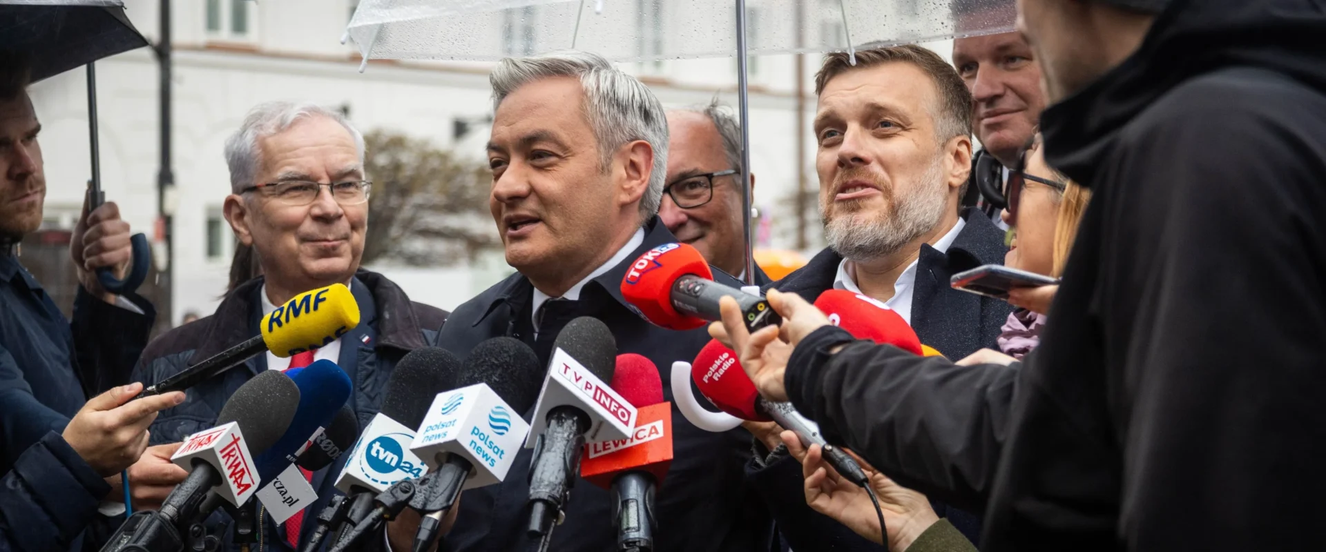 Mężczyzna w garniturze przemawia do licznych mikrofonów dziennikarzy podczas konferencji prasowej na zewnątrz, w deszczowy dzień, otoczony reporterami i parasolami.