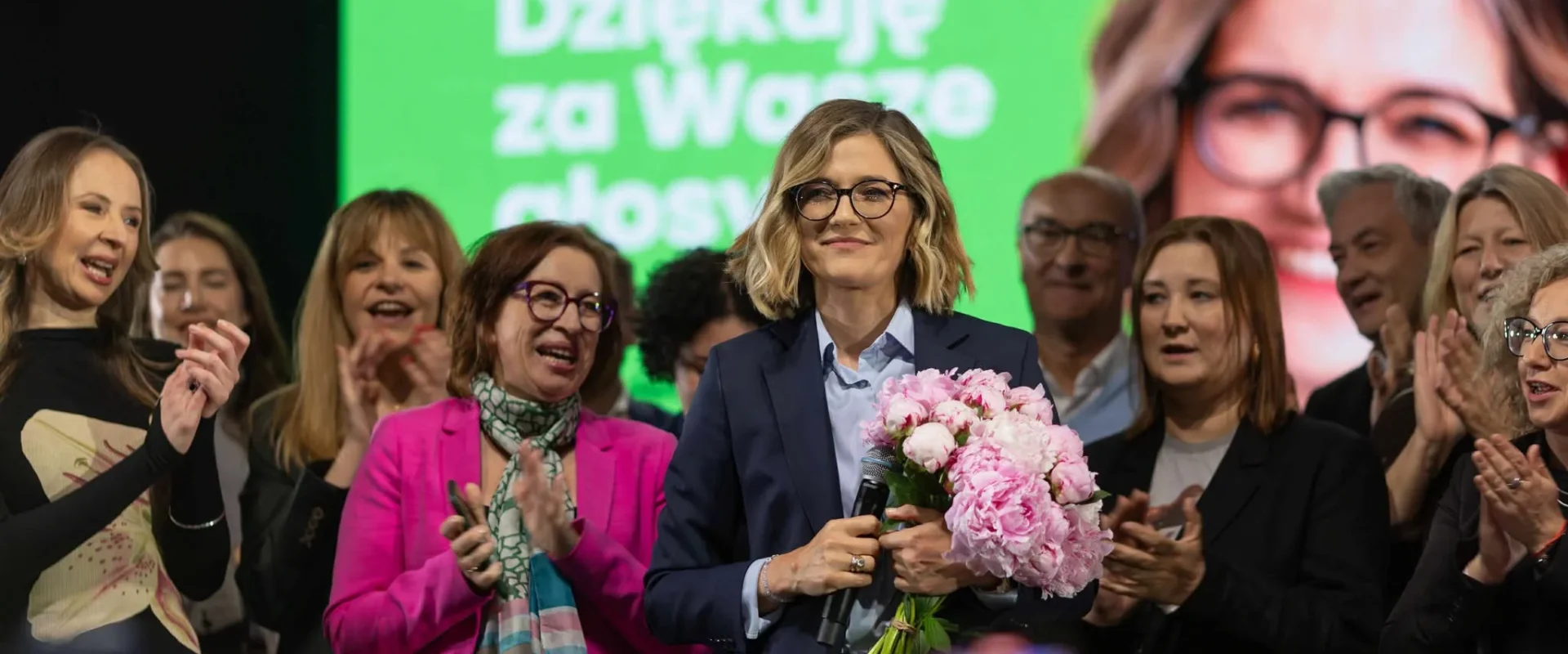 Na scenie stoi kobieta w granatowym garniturze, trzymająca mikrofon i bukiet różowych piwonii, otoczona wiwatującymi i klaskającymi ludźmi, a w tle widnieje jasny zielony ekran z napisem „Dziękuję za Wasze głosy”.