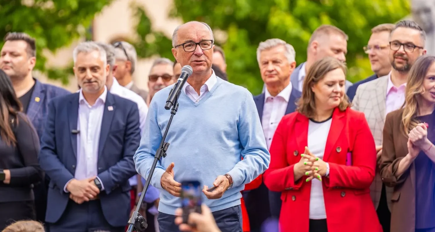 Mężczyzna w jasnoniebieskim swetrze przemawia do mikrofonu podczas konferencji w kontekście tragedii w Amazonii, otoczony grupą ludzi na tle zielonych drzew.