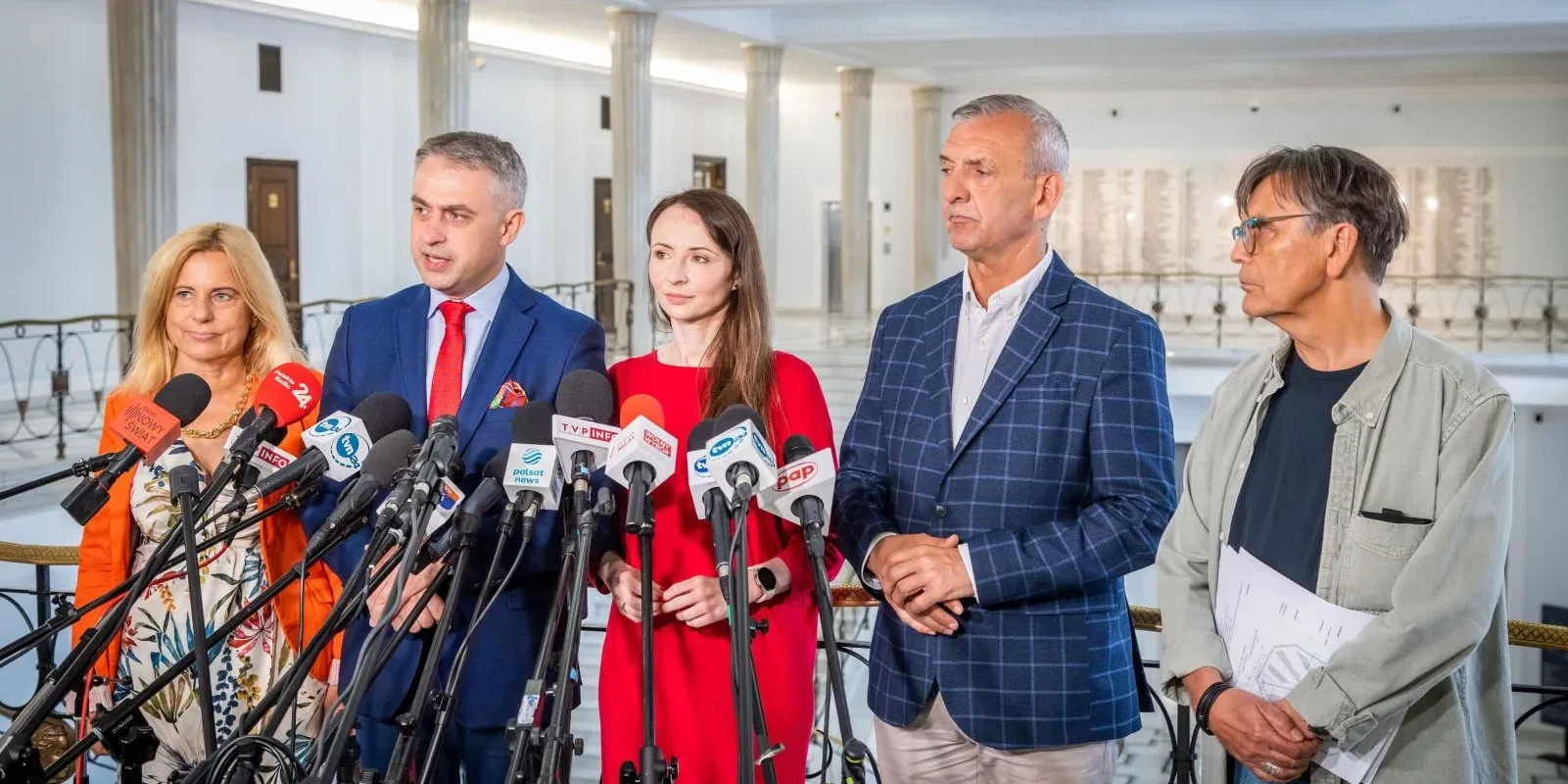 Na zdjęciu znajduje się grupa osób stojących przy mikrofonach w budynku publicznym, podczas konferencji prasowej.