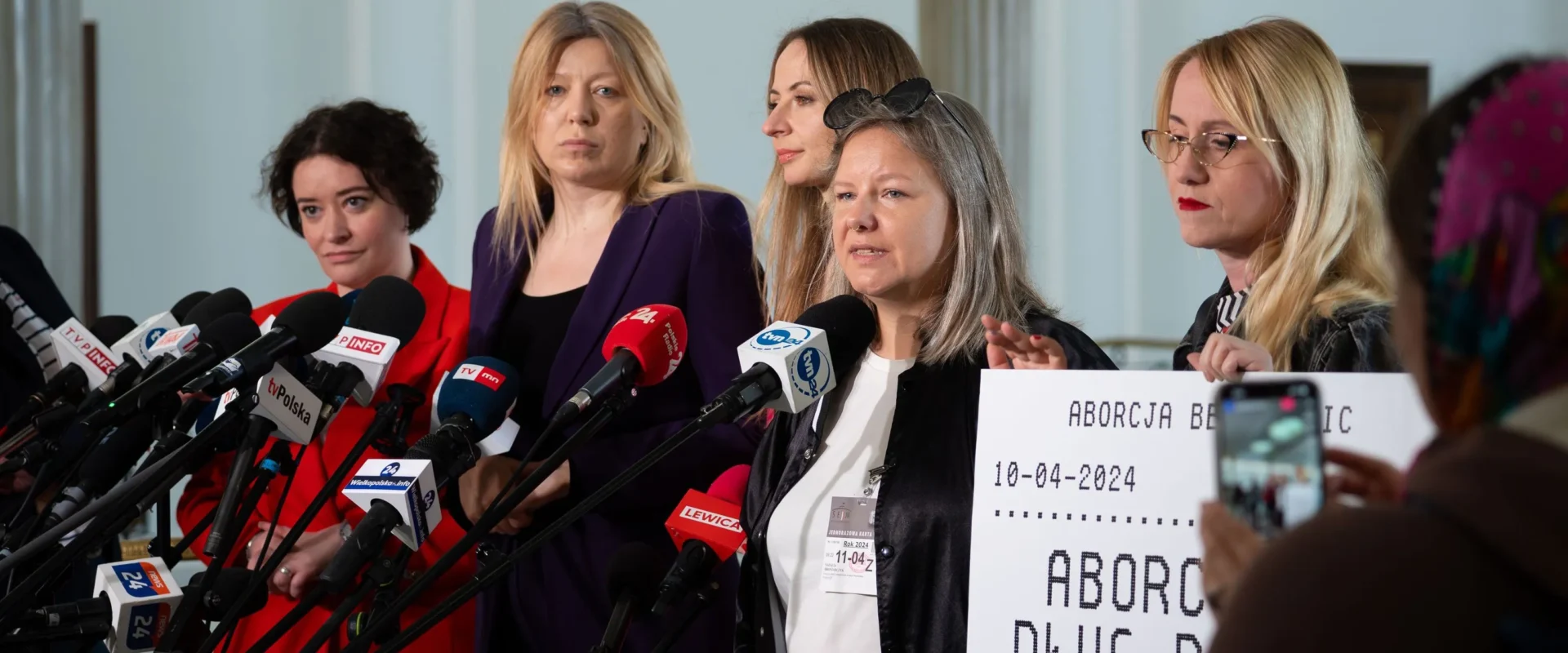 Grupa kobiet podczas konferencji prasowej w Sejmie stoi przed szeregiem mikrofonów, trzymając plakat dotyczący aborcji.