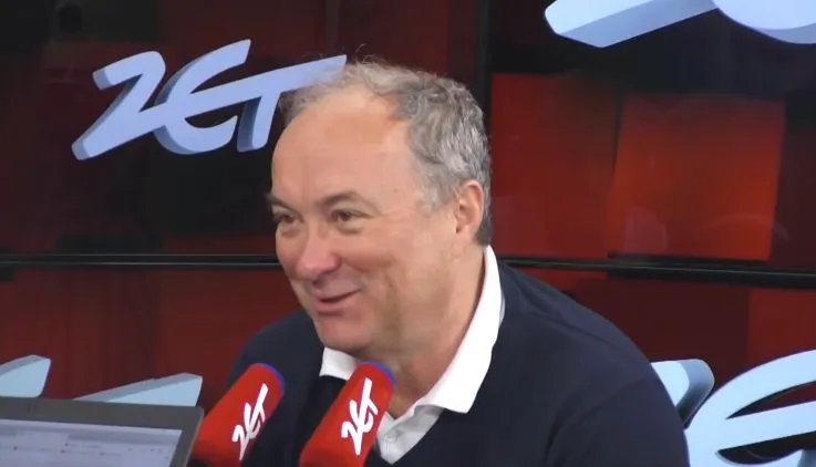 Mężczyzna w białej koszuli i granatowym swetrze siedzi w studiu radiowym i uśmiecha się, mówiąc do dwóch czerwonych mikrofonów z logo ZET.