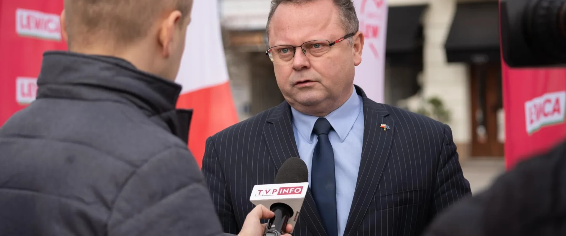 Mężczyzna w ciemnym, prążkowanym garniturze i okularach udziela wywiadu reporterowi TVP Info podczas konferencji prasowej; w tle widoczne są czerwone banery z logotypami.