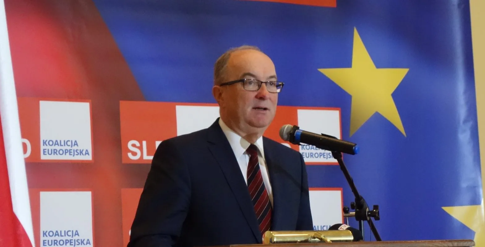 Mężczyzna w garniturze wygłasza przemówienie z podium podczas konferencji, a w tle widoczne są flaga Unii Europejskiej i banery Koalicji Europejskiej.