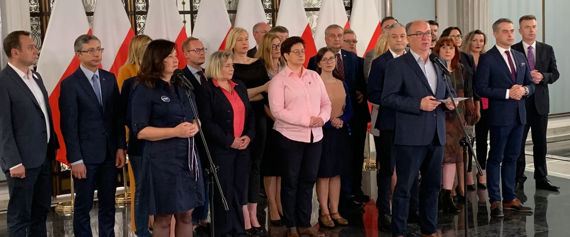 Grupa polityków stoi na tle flag Polski w eleganckiej sali, podczas gdy mężczyzna w garniturze przemawia do mikrofonu.