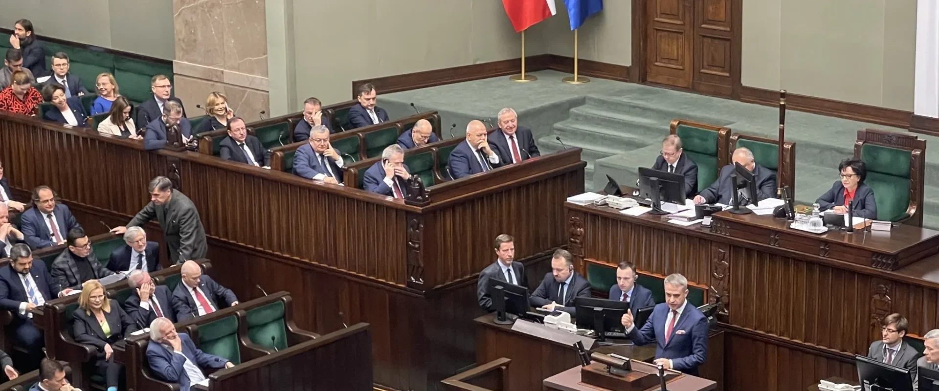 Na zdjęciu widoczna jest sala sejmowa, w której posłowie dyskutują na temat braku funduszy z KPO, a w centralnej części przemawia mężczyzna.