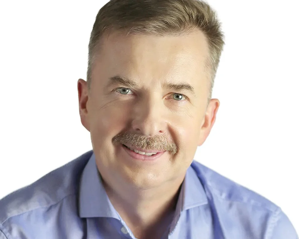 Portret uśmiechniętego mężczyzny z krótkimi włosami i wąsami w jasnoniebieskiej koszuli na białym tle.