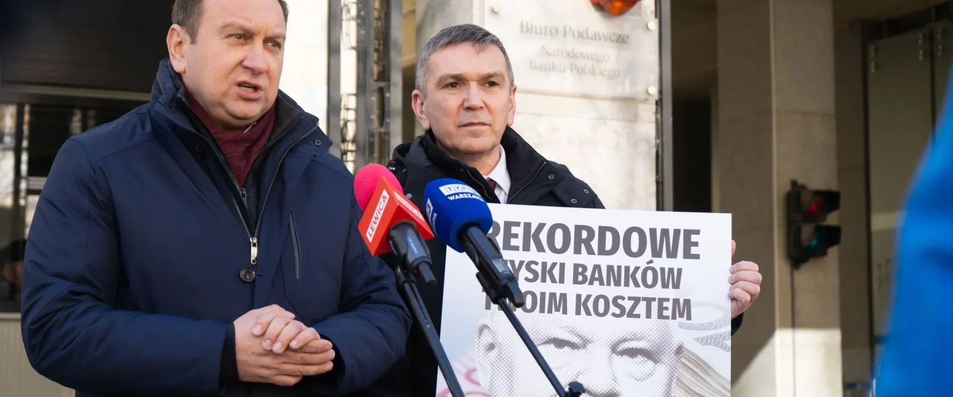 Dwóch mężczyzn na konferencji prasowej na zewnątrz, trzymają plakat o kosztach banków i przemawiają do mikrofonów, w kontekście apelu Lewicy o obniżenie stóp procentowych.