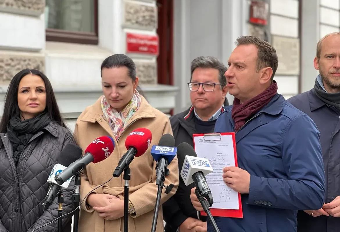 Grupa osób na konferencji prasowej na zewnątrz budynku; mężczyzna z teczką przemawia do zestawu mikrofonów, otoczony przez dwie kobiety i innych mężczyzn.