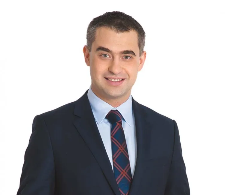 Mężczyzna w ciemnym garniturze, jasnej koszuli i kraciastej krawacie uśmiecha się na białym tle.