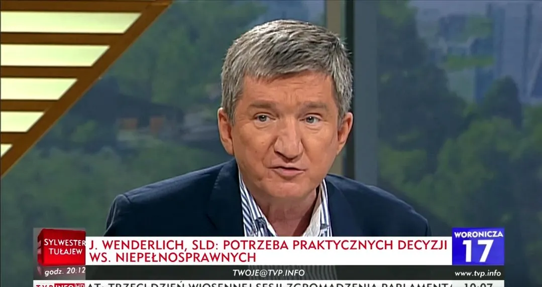 Mężczyzna w garniturze prowadzi program telewizyjny w studiu, a na dole ekranu przewija się pasek informacyjny o decyzjach ws. niepełnosprawnych.