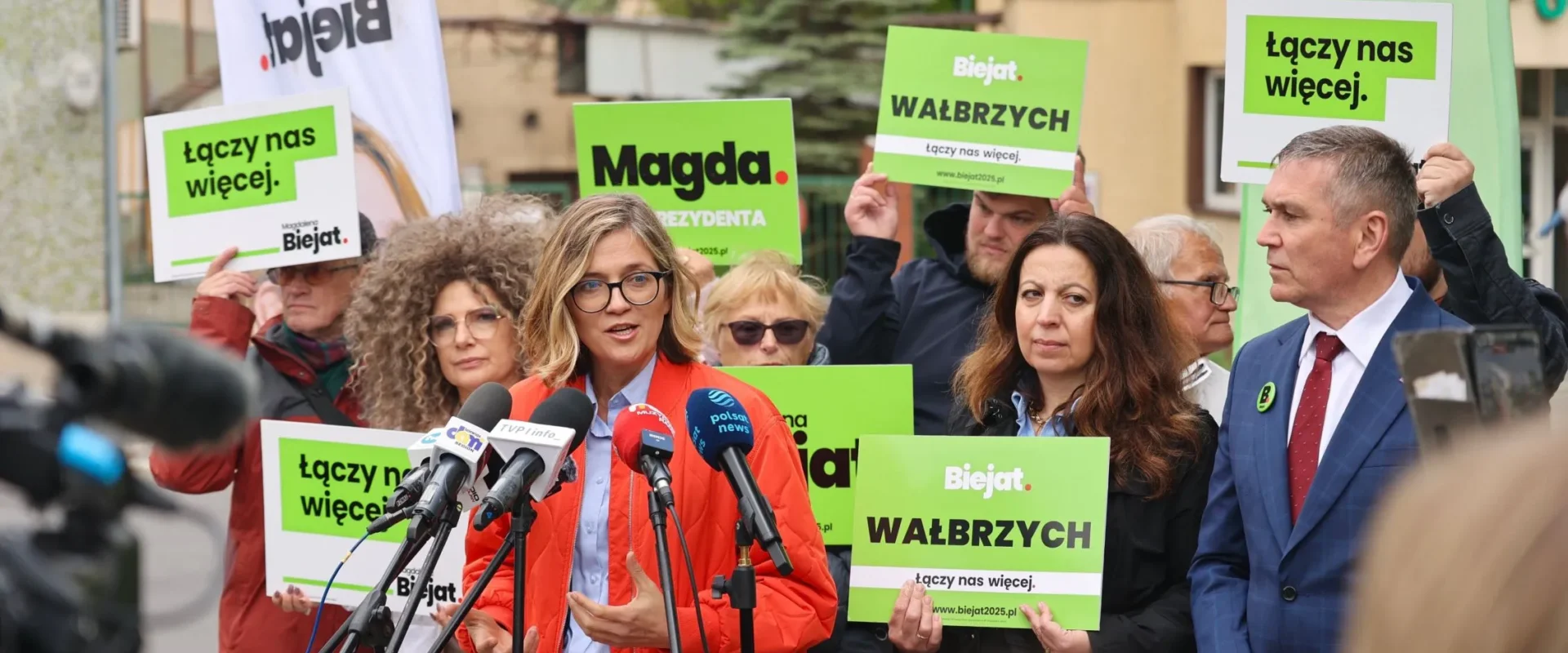 Grupa zwolenników na konferencji prasowej trzyma zielone plakaty z hasłem „Łączy nas więcej” i logiem Biejat, gdy kobieta w czerwonej kurtce przemawia do mikrofonów.