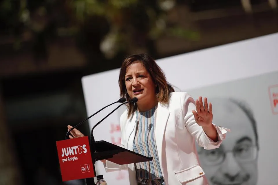 Polityczka w białej marynarce przemawia z podiumu, gestykulując prawą ręką, na tle baneru z napisem „JUNTOS por Aragón” i dużego plakatu z wizerunkiem twarzy.