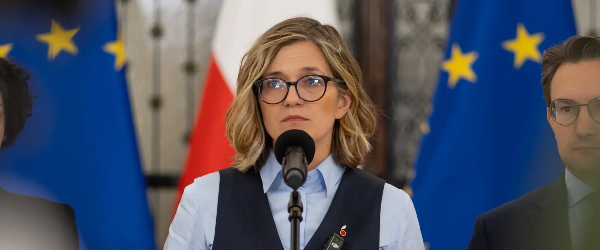 Kobieta w okularach przemawia do mikrofonu podczas konferencji prasowej, z flagami Unii Europejskiej i Polski w tle.