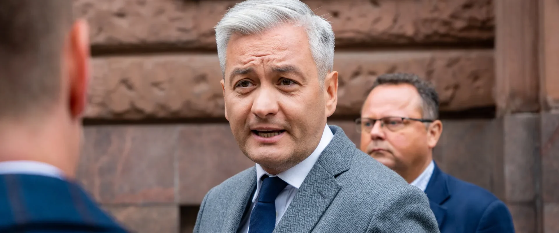Mężczyzna z siwymi włosami w szarym garniturze i granatowym krawacie rozmawia z rozmówcą na tle ceglanej fasady; w tle widoczny jest drugi mężczyzna w garniturze.