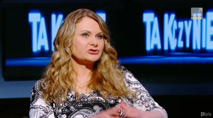 Kobieta z długimi, falującymi blond włosami gestykuluje i wypowiada się w studiu telewizyjnym, a za nią widać rozmyty, podświetlony napis programu.