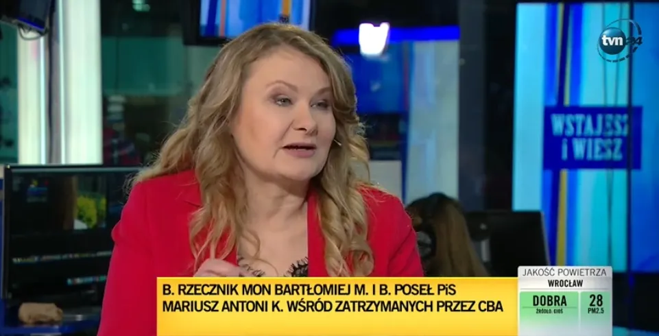 Kobieta z blond włosami w czerwonej marynarce siedzi w studiu telewizyjnym podczas programu informacyjnego, a na dole ekranu widnieje żółty pasek z nagłówkiem i logo TVN w tle.