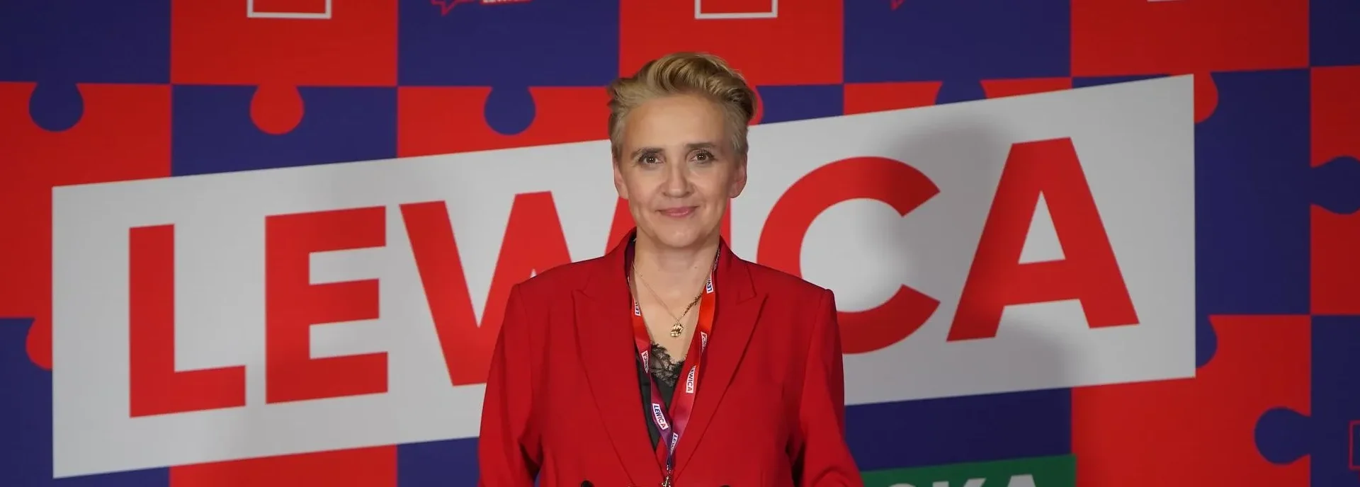 Polityczka w czerwonej marynarce stoi na tle kolorowych puzzli z dużym napisem LEWICA, podczas konferencji o zmianach w mediach publicznych.