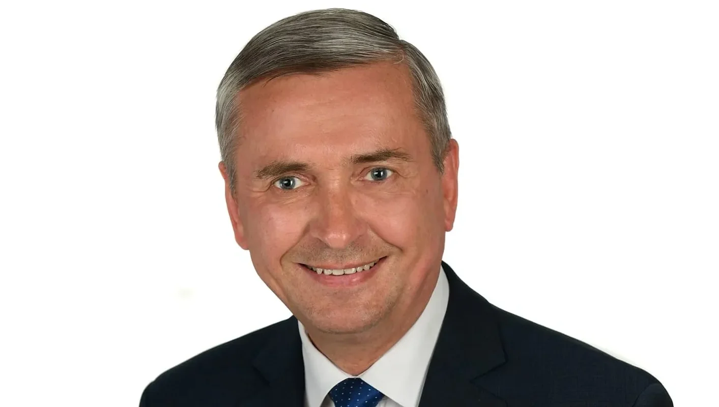 Portret mężczyzny w garniturze i niebieskim krawacie w białe kropki na białym tle, uśmiechnięty.