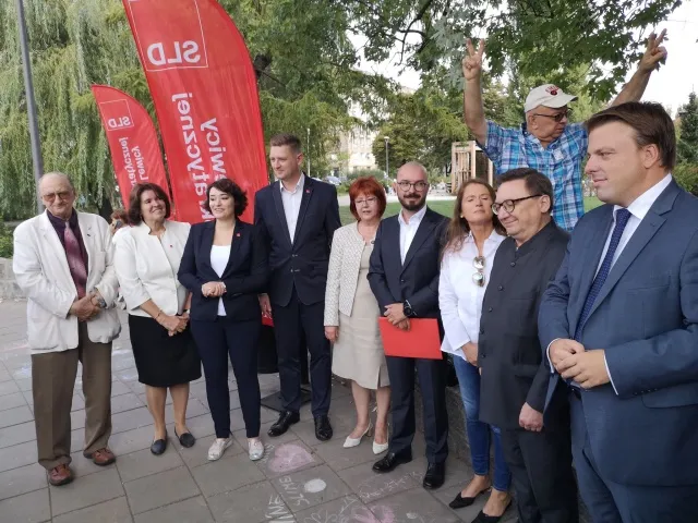 Grupa liderów komitetu SLD Lewica Razem stoi na zewnątrz, ubrani w stroje formalne, trzymając czerwone teczki, w tle widoczne czerwone flagi z napisem SLD.