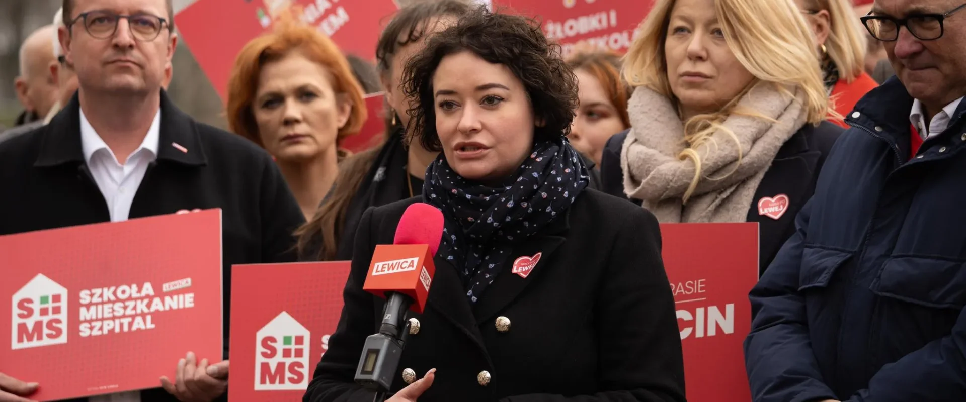 Kobieta przemawia na wiecu Lewicy, otoczona innymi uczestnikami trzymającymi czerwone plakaty z hasłami i logo partii.